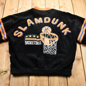 May include: Black suede bomber jacket with orange and blue striped sleeves. The back of the jacket features an embroidered patch with the words "Slam Dunk" and a basketball going through a hoop.