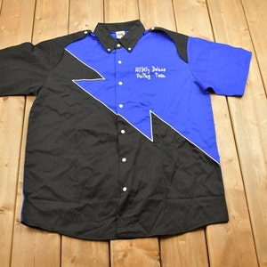 Può includere: Una camicia a maniche corte con bottoni nera e blu con un design a forma di fulmine bianco. La camicia ha il testo "Artley Deluxe Pulling Team" ricamato sul davanti.