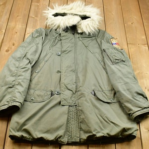 Puede incluir: Una parka verde con capucha forrada de piel. La chaqueta tiene cierre de cremallera, dos bolsillos con solapa y un parche en la manga izquierda.