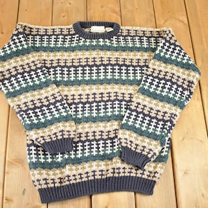 May include: A patterned crew neck sweater in navy, green, beige, and white. The sweater features a ribbed collar, cuffs, and hem. The knitwear has a geometric design. The sweater is made of a knitted fabric.