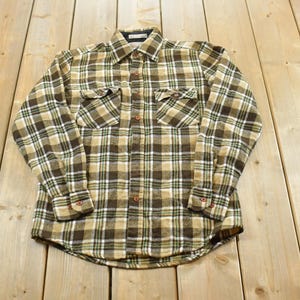 May include: A long-sleeve button-down shirt featuring a plaid pattern in shades of brown, tan, and white. The shirt includes a standard collar, a full-length front button closure, two chest pockets with buttoned flaps, and buttoned cuffs on the sleeves.