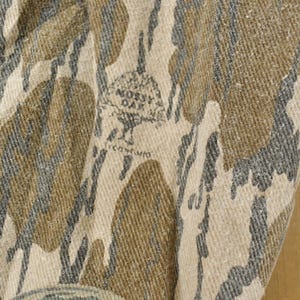 May include: Close-up of a fabric with a camouflage pattern in shades of brown, gray, and beige. The fabric has the "Mossy Oak" logo and text. The pattern is a blend of organic shapes.