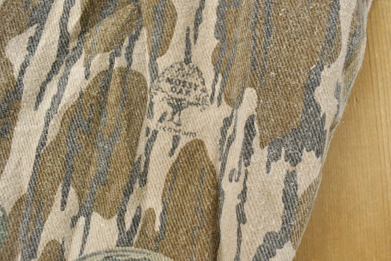 May include: Close-up of a fabric with a camouflage pattern in shades of brown, gray, and beige. The fabric has the "Mossy Oak" logo and text. The pattern is a blend of organic shapes.