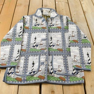 May include: A light blue jacket featuring a repeating pattern of lighthouses, sailboats, and birds. The jacket has a button-up front, two pockets, and a light blue trim. The jacket is made of a woven fabric.