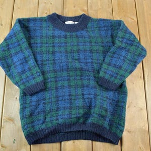 Könnte beinhalten: Ein blau-grüner karierter Pullover mit Rundhalsausschnitt und langen Ärmeln. Der Pullover hat einen gerippten Saum und Bündchen. Das Karomuster ist eine Mischung aus blauen und grünen Farbtönen. Der Pullover ist aus weichem, strukturiertem Material.