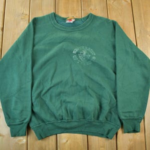 Puede incluir: Una sudadera verde vintage con cuello redondo y mangas largas. La sudadera presenta un gráfico descolorido en el pecho. La prenda está hecha de un material suave y tiene puños y dobladillo acanalados.