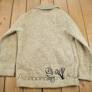 May include: A light gray knit cardigan with a collar. The back of the sweater features a black embroidered design of an astronaut, a tree, and a wavy line. The sweater is made of a textured knit fabric.