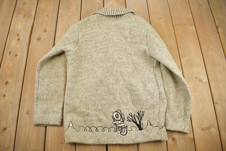 May include: A light gray knit cardigan with a collar. The back of the sweater features a black embroidered design of an astronaut, a tree, and a wavy line. The sweater is made of a textured knit fabric.
