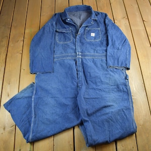 Könnte beinhalten: Ein blauer Jeans-Overall mit langen Ärmeln, einem Button-Down-Kragen und mehreren Taschen. Das Kleidungsstück im Workwear-Stil liegt flach auf einer Holzoberfläche. Der Overall hat ein kleines weißes Etikett auf der Brust.