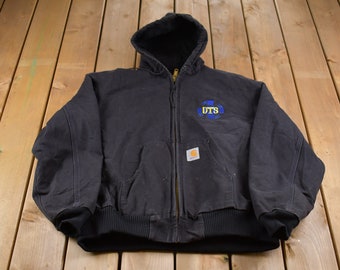 Puede incluir: Chaqueta negra con capucha y cremallera, con un logotipo DTS azul y amarillo en el pecho. La chaqueta tiene un dobladillo y puños de canalé negros, y una etiqueta Carhartt. La chaqueta se muestra sobre una superficie de madera.