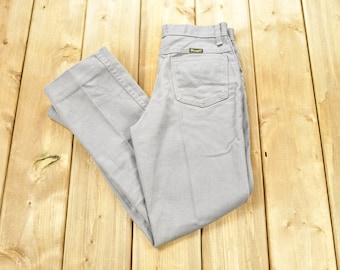 Può includere: Un paio di pantaloni grigi chiari piegati a metà su una superficie di legno. I pantaloni hanno un'etichetta Wrangler sulla tasca posteriore.