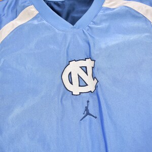 May include: A light blue sleeveless basketball jersey with white accents and a white "UNC" logo with a blue outline. A black and white silhouette of a basketball player is below the logo.