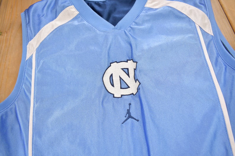 May include: A light blue sleeveless basketball jersey with white accents and a white "UNC" logo with a blue outline. A black and white silhouette of a basketball player is below the logo.