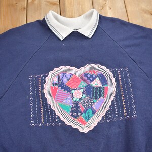 May include: A navy blue sweatshirt with a white collar and a large embroidered heart design. The heart is made up of colorful patchwork fabric.