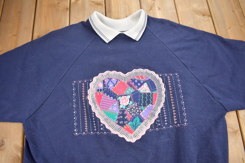 May include: A navy blue sweatshirt with a white collar and a large embroidered heart design. The heart is made up of colorful patchwork fabric.