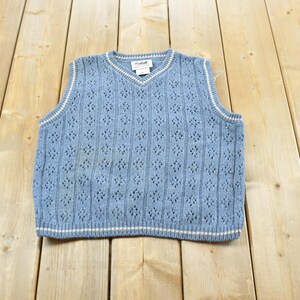 May include: A light blue sleeveless sweater vest featuring a V-neckline with white and blue striped trim. The knit fabric displays a vertical pattern of openwork diamond shapes and ribbed columns. A white brand label is visible at the inner neckline.