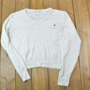 Könnte beinhalten: Ein weißer Langarmpullover mit V-Ausschnitt und einem kleinen roten Emblem auf der Brust. Der Pullover ist aus einem weichen Material gefertigt und hat einen leicht verkürzten Saum. Das Kleidungsstück wird auf einer Holzoberfläche präsentiert.