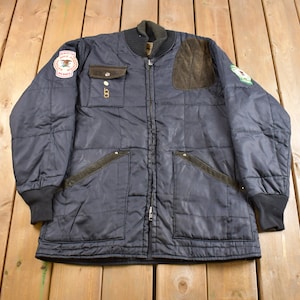 Puede incluir: Una chaqueta acolchada azul marino con parches de gamuza marrón y cierre de cremallera. La chaqueta tiene dos bolsillos grandes en la parte delantera y un bolsillo más pequeño en el pecho izquierdo. La chaqueta tiene un parche en el pecho izquierdo con el texto "Golden Eagle" y un parche en el pecho derecho con el texto "Golden Eagle".