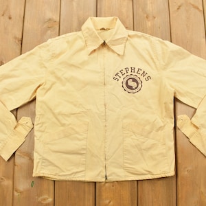 May include: A yellow vintage jacket with a zipper closure and two pockets. The jacket has a brown embroidered logo that says "Stephens" with a design inside the circle.