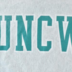 Puede incluir: Una sudadera gris con el texto "UNCW" en azul turquesa impreso en la parte delantera.