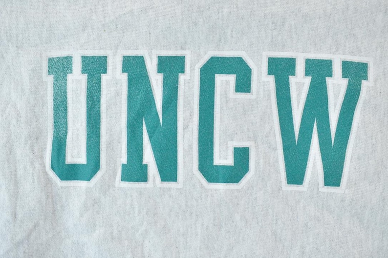 Puede incluir: Una sudadera gris con el texto "UNCW" en azul turquesa impreso en la parte delantera.