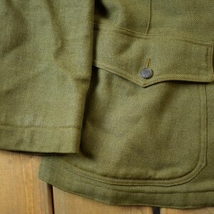 May include: A green wool jacket with a large flap pocket on the right side. The pocket has a button closure. The jacket has a textured weave.