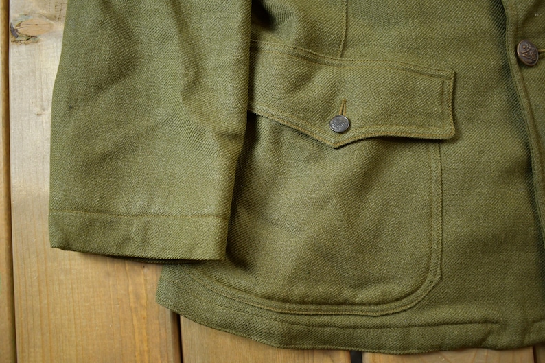 May include: A green wool jacket with a large flap pocket on the right side. The pocket has a button closure. The jacket has a textured weave.