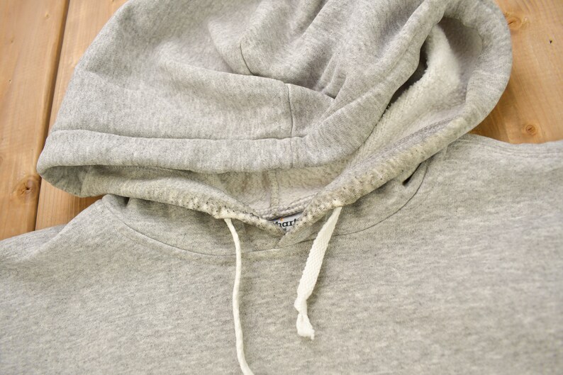 Puede incluir: Sudadera gris con capucha y cord&oacute;n blanco. La sudadera tiene una peque&ntilde;a etiqueta que dice "Champion" en el interior de la capucha.