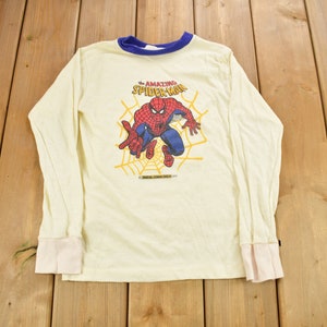 May include: A white long-sleeved shirt with a blue and white striped collar. The shirt features a graphic of Spider-Man in a web, with the text "The Amazing Spider-Man" above him.