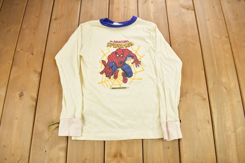 May include: A white long-sleeved shirt with a blue and white striped collar. The shirt features a graphic of Spider-Man in a web, with the text "The Amazing Spider-Man" above him.