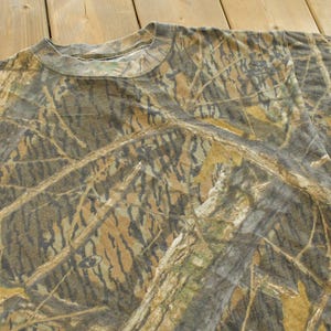 Puede incluir: Camiseta de manga corta con estampado de camuflaje en tonos marr&oacute;n, verde y beige. La tela tiene un aspecto texturizado y la camiseta tiene cuello redondo. El dise&ntilde;o general evoca un entorno forestal.