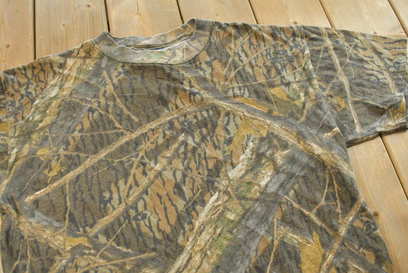 Puede incluir: Camiseta de manga corta con estampado de camuflaje en tonos marr&oacute;n, verde y beige. La tela tiene un aspecto texturizado y la camiseta tiene cuello redondo. El dise&ntilde;o general evoca un entorno forestal.