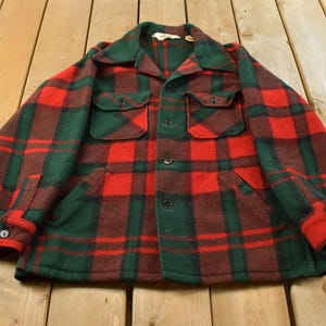 Pode incluir: Uma camisa de manga comprida com botões e um padrão xadrez em vermelho e verde escuro. A camisa inclui um colarinho pontiagudo, dois bolsos no peito com aba e botão, dois bolsos laterais inferiores e botões pretos na frente e nos punhos.