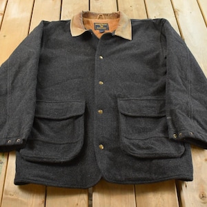 Puede incluir: Chaqueta gris oscuro con cuello marrón y botones dorados. La chaqueta tiene dos bolsillos delanteros grandes y está hecha de un material grueso y cálido. La chaqueta está sobre una superficie de madera.