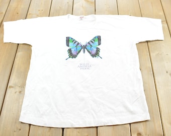 May include: A white short-sleeve t-shirt featuring a graphic of a butterfly with blue, green, and black wings. Below the butterfly, the text "WILD ANIMAL PARK" is printed in a simple font. The shirt has a crew neckline and a standard fit.