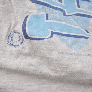May include: A gray t-shirt with a blue and white graphic print. The graphic is a stylized letter "L" with a blue and white pattern. The shirt has a small, circular tag with the text "Officially Licensed Collegiate Products" and a logo. The bottom of the shirt has the text "1993 Rah-Rah Sales Inc. 11301".