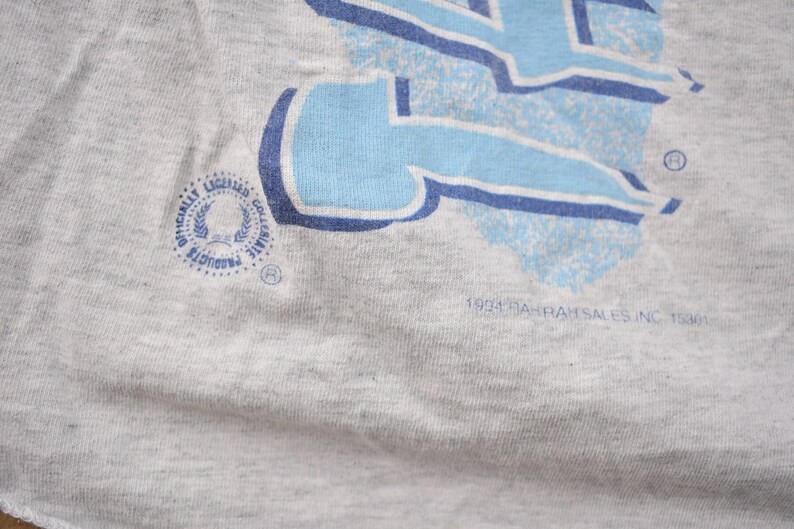 May include: A gray t-shirt with a blue and white graphic print. The graphic is a stylized letter "L" with a blue and white pattern. The shirt has a small, circular tag with the text "Officially Licensed Collegiate Products" and a logo. The bottom of the shirt has the text "1993 Rah-Rah Sales Inc. 11301".