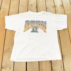 May include: White t-shirt with the text "DOOM II" in a multi-colored, 3D-style graphic. The shirt is made of a soft material and is laid flat on a wooden surface. The t-shirt is a vintage item.