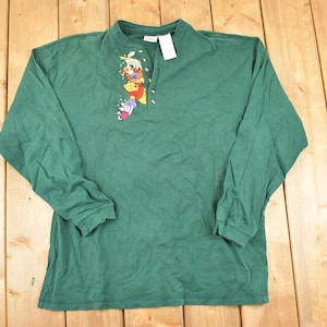 May include: A green long-sleeve shirt featuring a small Winnie the Pooh graphic on the chest. The shirt has a mock neck and a partial zipper. A white tag is attached to the collar. The shirt is made of a soft material.