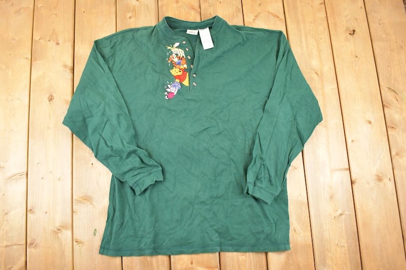 May include: A green long-sleeve shirt featuring a small Winnie the Pooh graphic on the chest. The shirt has a mock neck and a partial zipper. A white tag is attached to the collar. The shirt is made of a soft material.