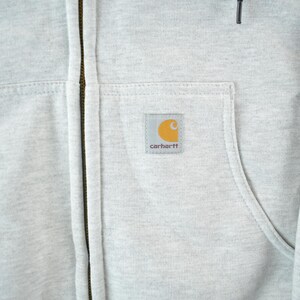 Puede incluir: Primer plano de una sudadera con capucha gris claro con cremallera. La sudadera con capucha tiene un bolsillo delantero y un peque&ntilde;o parche cuadrado con el logotipo de Carhartt. La cremallera es de color marr&oacute;n oscuro. La tela parece ser un material suave y duradero.