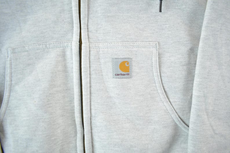 Puede incluir: Primer plano de una sudadera con capucha gris claro con cremallera. La sudadera con capucha tiene un bolsillo delantero y un peque&ntilde;o parche cuadrado con el logotipo de Carhartt. La cremallera es de color marr&oacute;n oscuro. La tela parece ser un material suave y duradero.