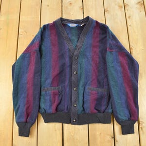 Puede incluir: Un c&aacute;rdigan vintage con botones y cuello en V. El su&eacute;ter presenta rayas verticales en tonos azules, verdes y burdeos. Tiene dos bolsillos delanteros y un dobladillo y pu&ntilde;os de canal&eacute; negros. La prenda est&aacute; sobre una superficie de madera.