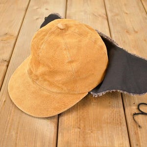 Può includere: Un cappello color marrone chiaro con paraorecchie e una visiera curva. Il cappello ha un aspetto strutturato e una fodera grigio scuro sui paraorecchie. Cordini neri pendono dai paraorecchie. Il cappello è su una superficie di legno.