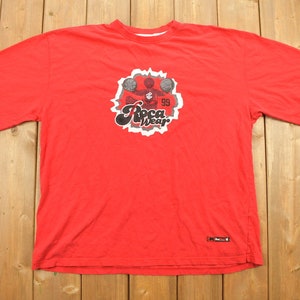 Puede incluir: Camiseta roja con un gráfico blanco de un jugador de baloncesto que lleva una camiseta de Rocawear y sostiene un balón de baloncesto. El gráfico también incluye el texto "Rocawear" y "99".