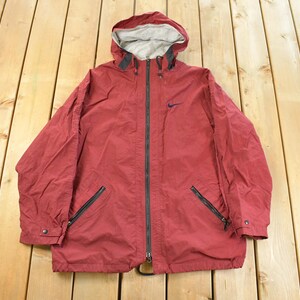 May include: A red Nike hooded jacket with a full-length zipper, two front pockets, and the Nike logo on the chest. The jacket has a gray interior lining and black zippers. The jacket is laid flat on a wooden surface.