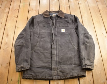 Könnte beinhalten: Eine dunkelbraune Carhartt Arbeitsjacke mit einem Cordkragen und einem Patch-Logo auf der Brust. Die Jacke hat zwei große Taschen vorne und zwei kleinere Taschen auf der Brust.