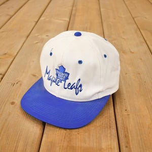 Può includere: Un cappellino da baseball bianco e blu con la scritta "Toronto Maple Leafs" in blu. Il cappellino ha una visiera blu e un logo a forma di foglia d'acero blu. Il cappello è su una superficie di legno.
