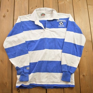 Könnte beinhalten: Ein blaues und weiß gestreiftes Langarm-Rugby-Shirt mit weißem Kragen und einem kleinen gestickten Logo mit der Aufschrift "Shaker Soccer".