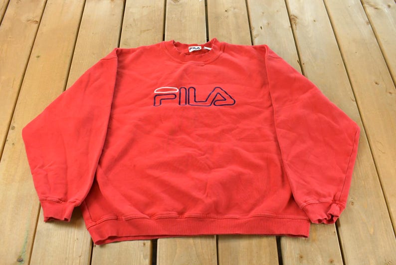 Puede incluir: Sudadera roja de FILA con el nombre de la marca bordado en blanco y azul marino en el pecho. La parte superior de cuello redondo y manga larga es de material suave.